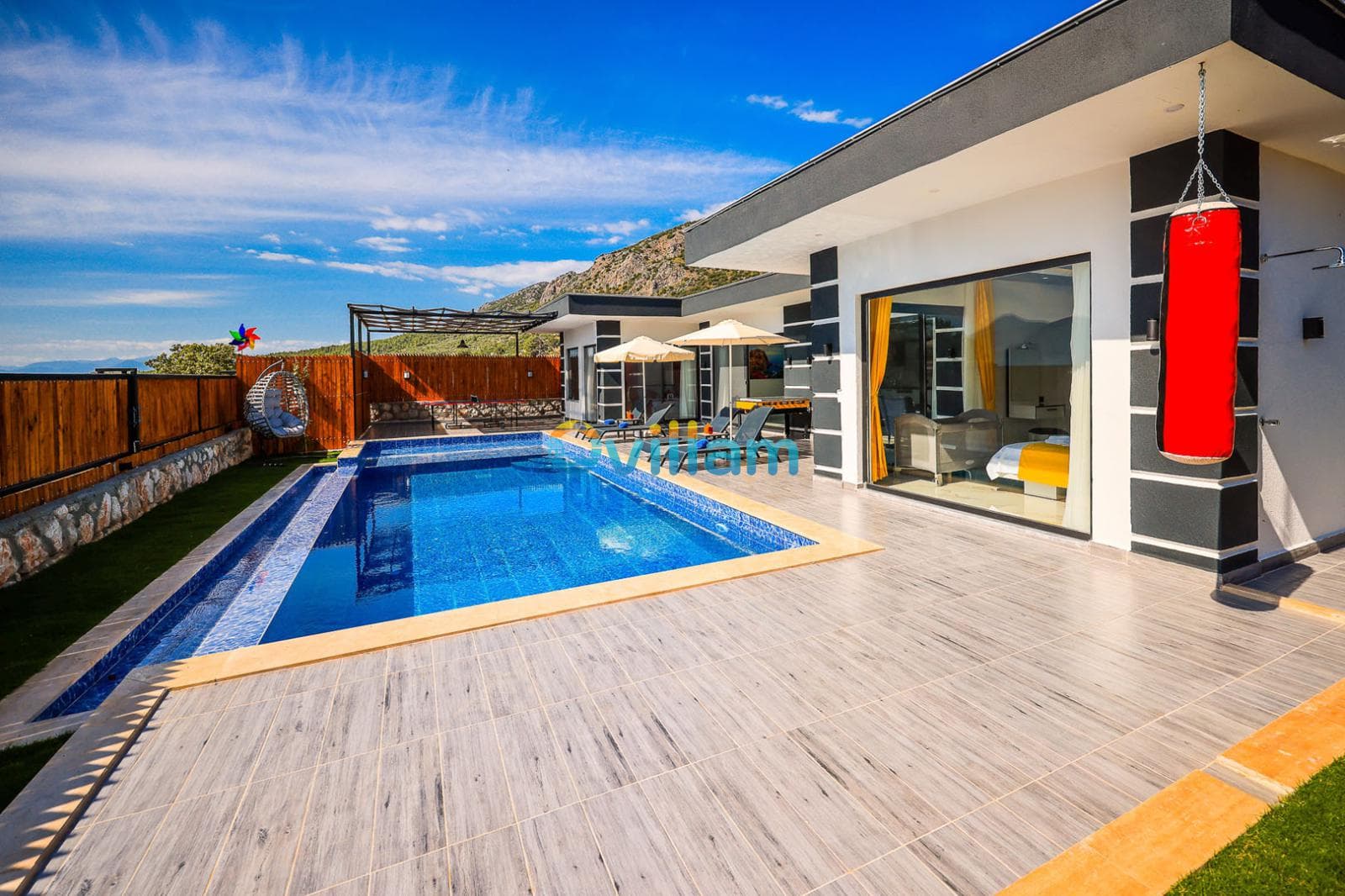 Açık havuzlu villanın terasında şezlonglar, güneş şemsiyeleri ve kırmızı bir boks torbası bulunuyor.