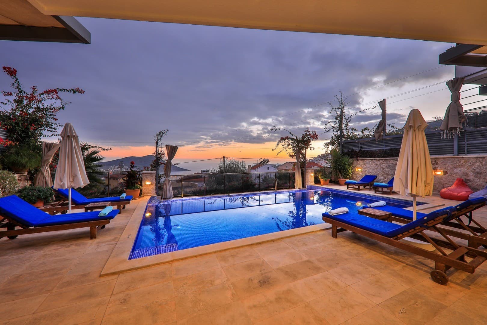 Gün batımında havuz başında şezlonglar ve güneş şemsiyeleri bulunan taş zeminli villa terası.