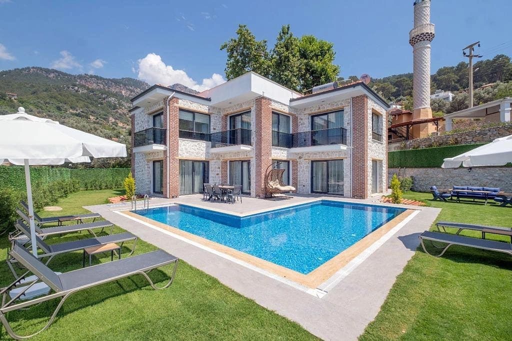 Taş ve tuğla detaylı iki katlı villa, havuz ve şezlonglar.