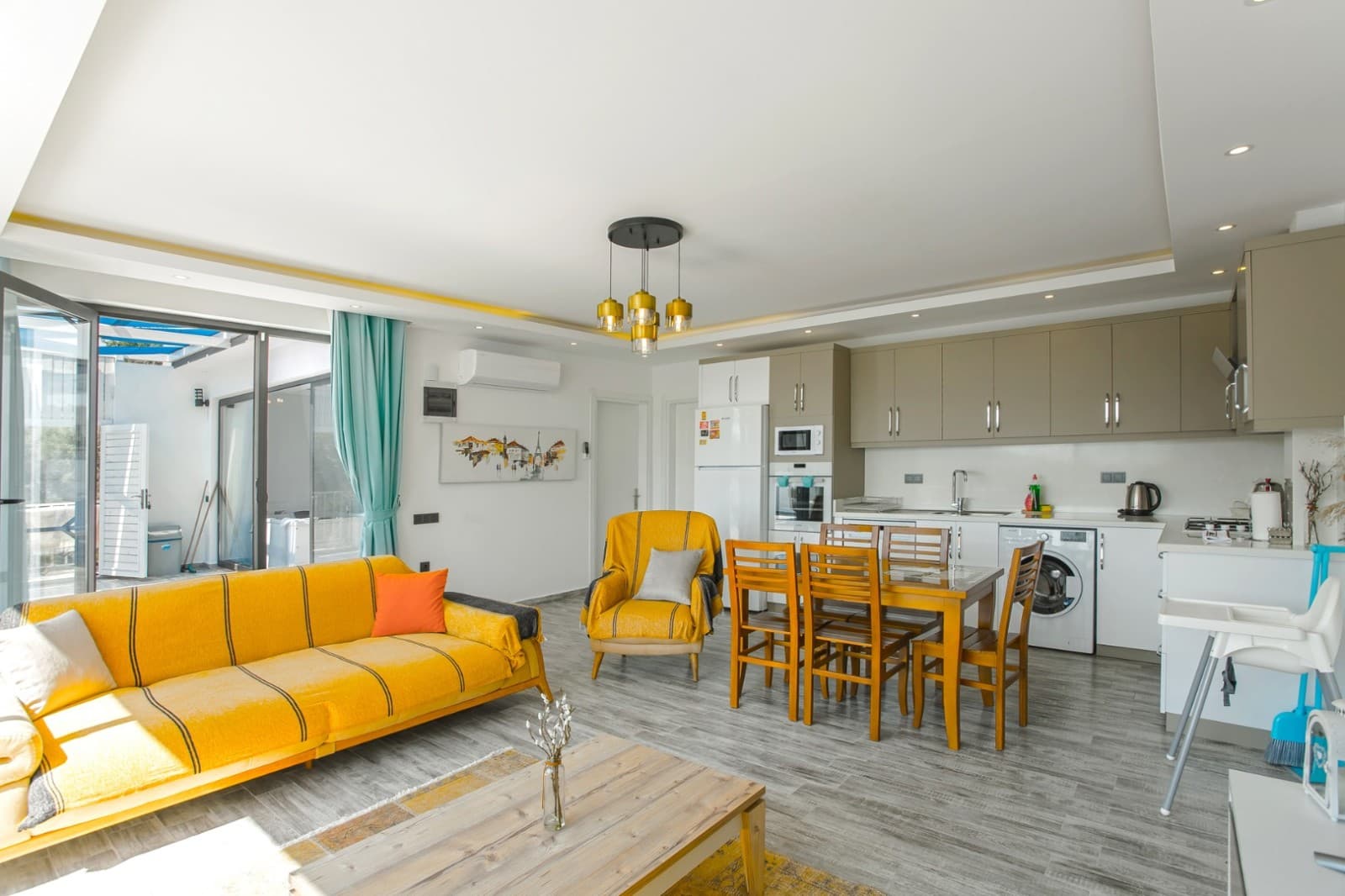 Sarı L koltuk, yemek masası ve açık mutfak bulunan aydınlık salon.