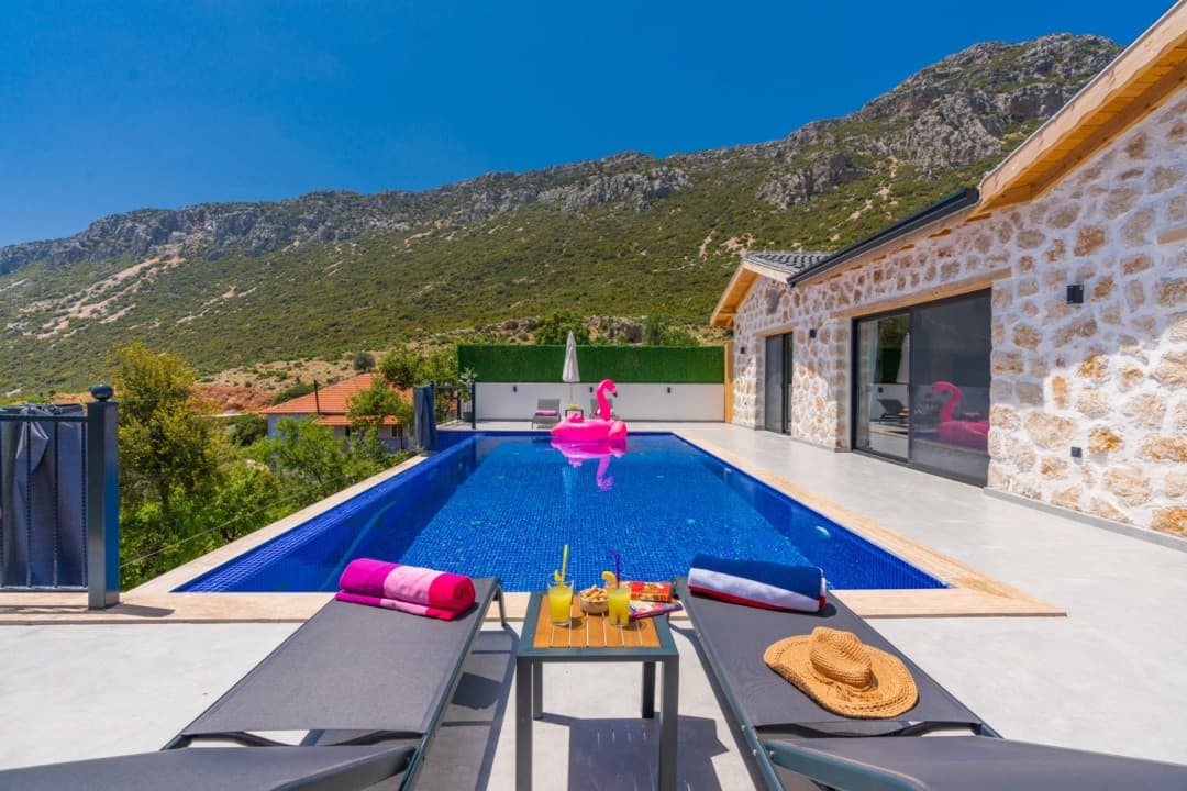 Ön planda şezlonglar, ortada havuz ve arka planda dağ manzarası bulunan taş duvarlı villa.