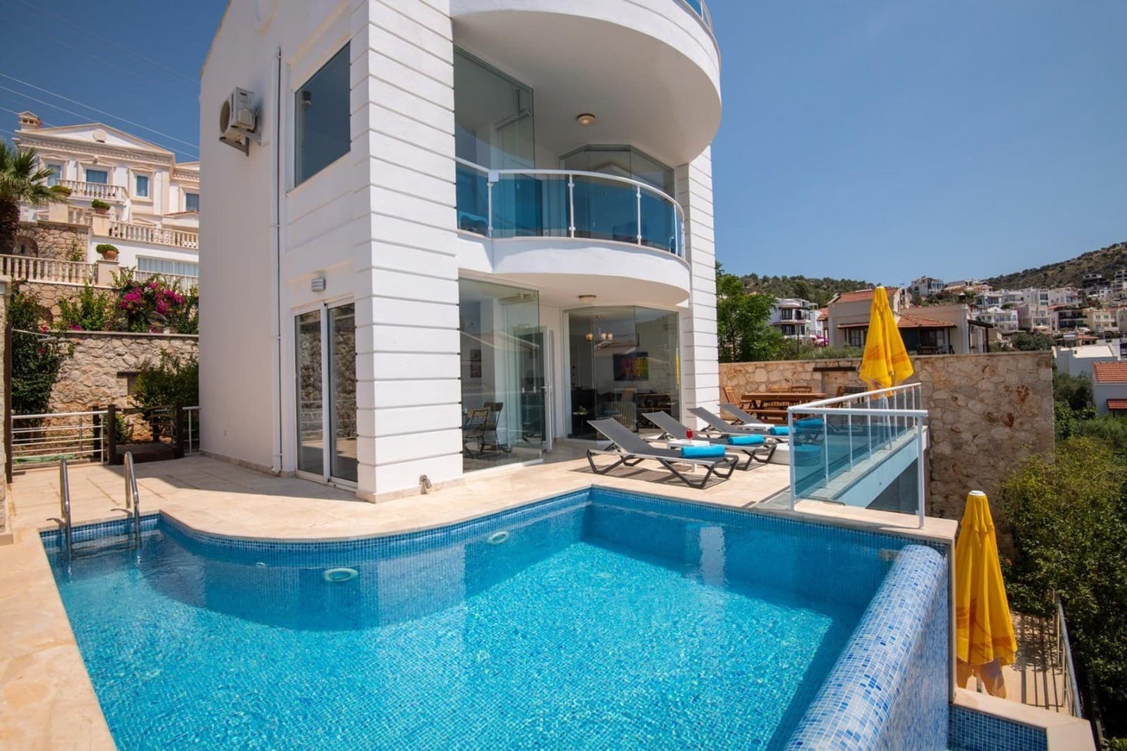 Önünde özel havuzu, şezlongları ve sarı güneş şemsiyeleri bulunan beyaz dış cepheli iki katlı villa.