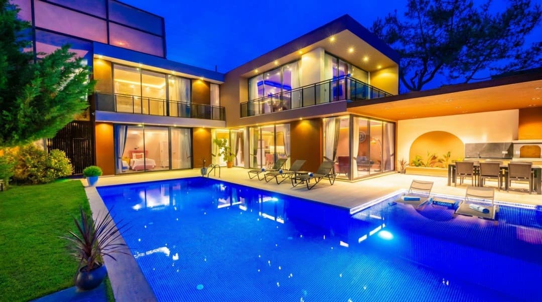 Akşam vakti aydınlatılmış modern villanın önünde havuz, şezlonglar ve yemek masası yer alıyor.