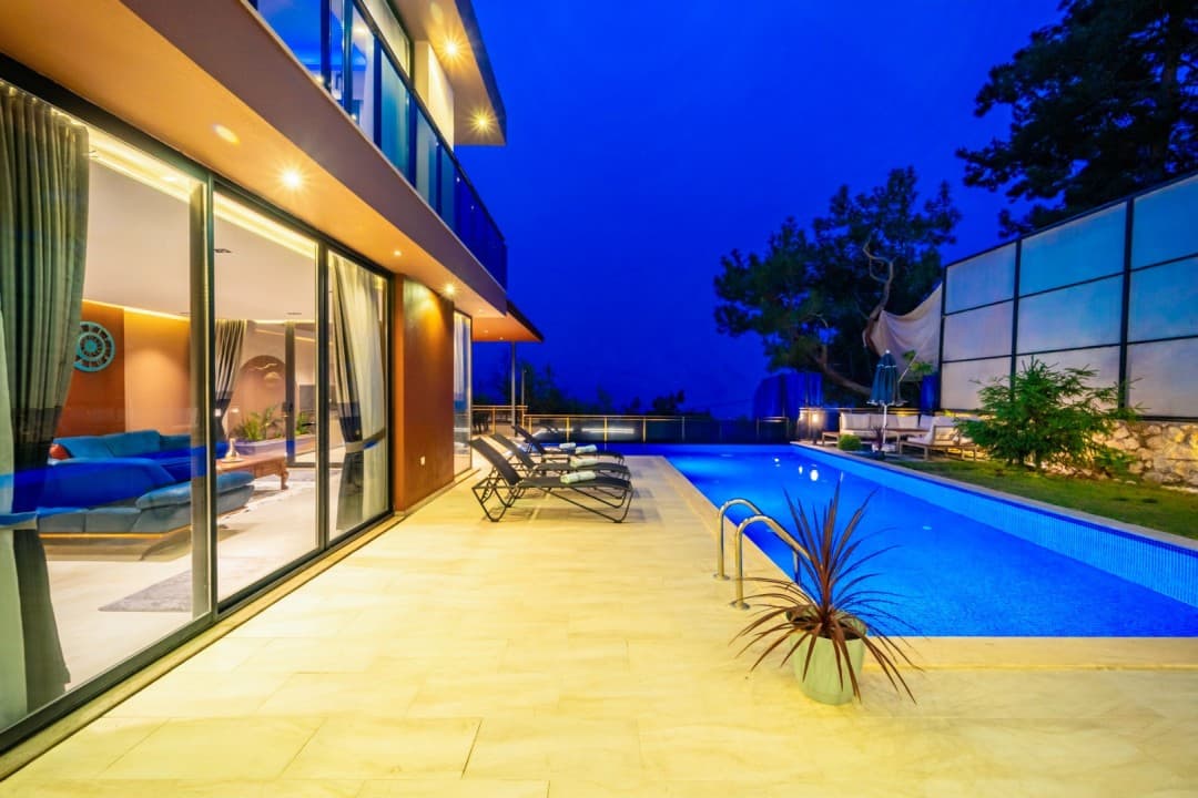 Gece aydınlatmalı havuz kenarında şezlonglar ve arkada villa.