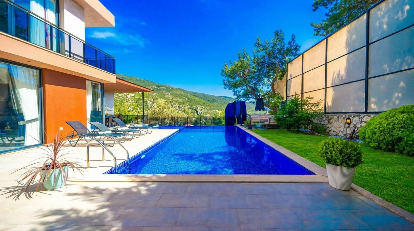Açık mavi havuz ve şezlongların bulunduğu, dağ manzaralı geniş villa bahçesi.