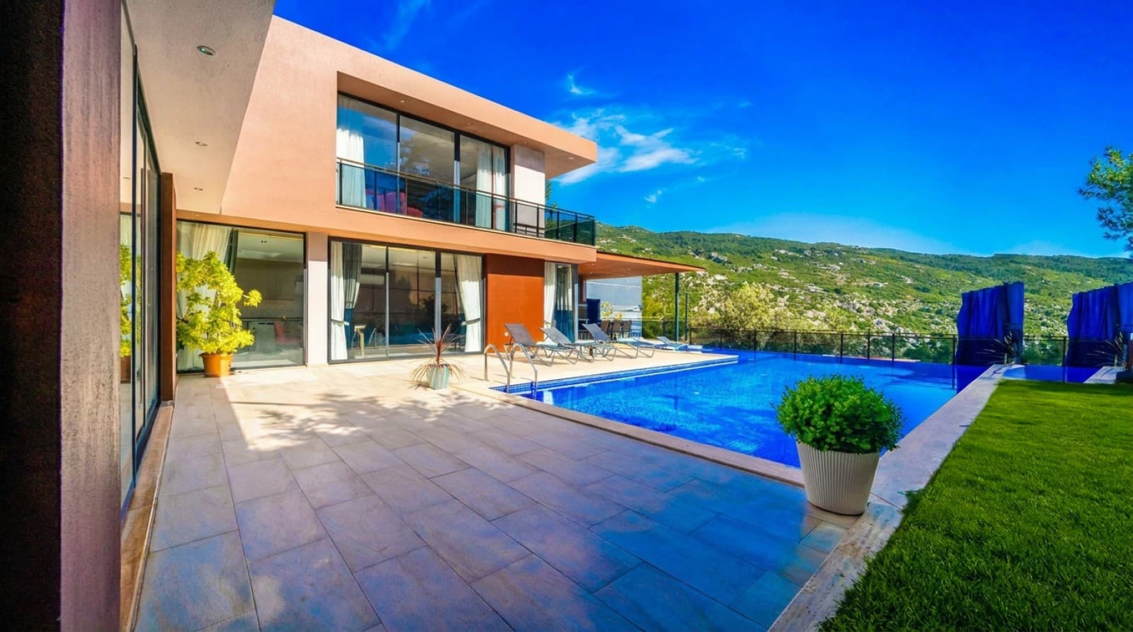 Geniş terasta yüzme havuzu, şezlonglar ve dağ manzaralı iki katlı modern villa dış cephesi.