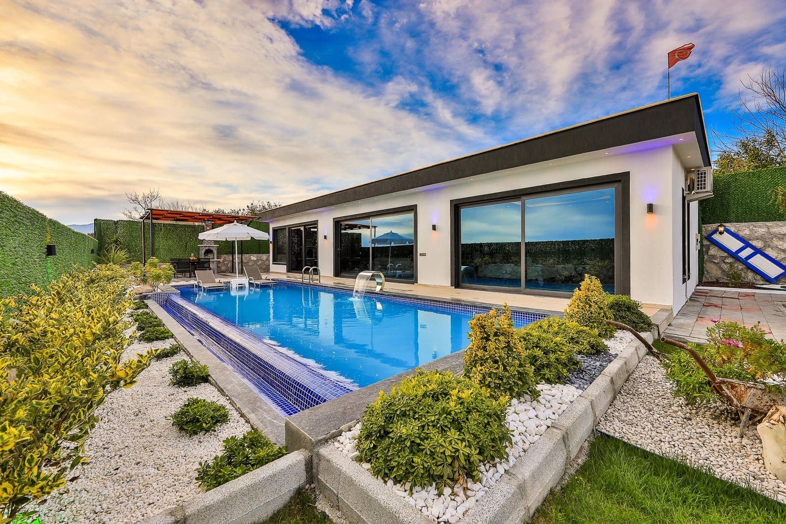 Özel yüzme havuzu, şezlongları ve peyzajlı bahçesi bulunan modern tek katlı villa dış cephesi.
