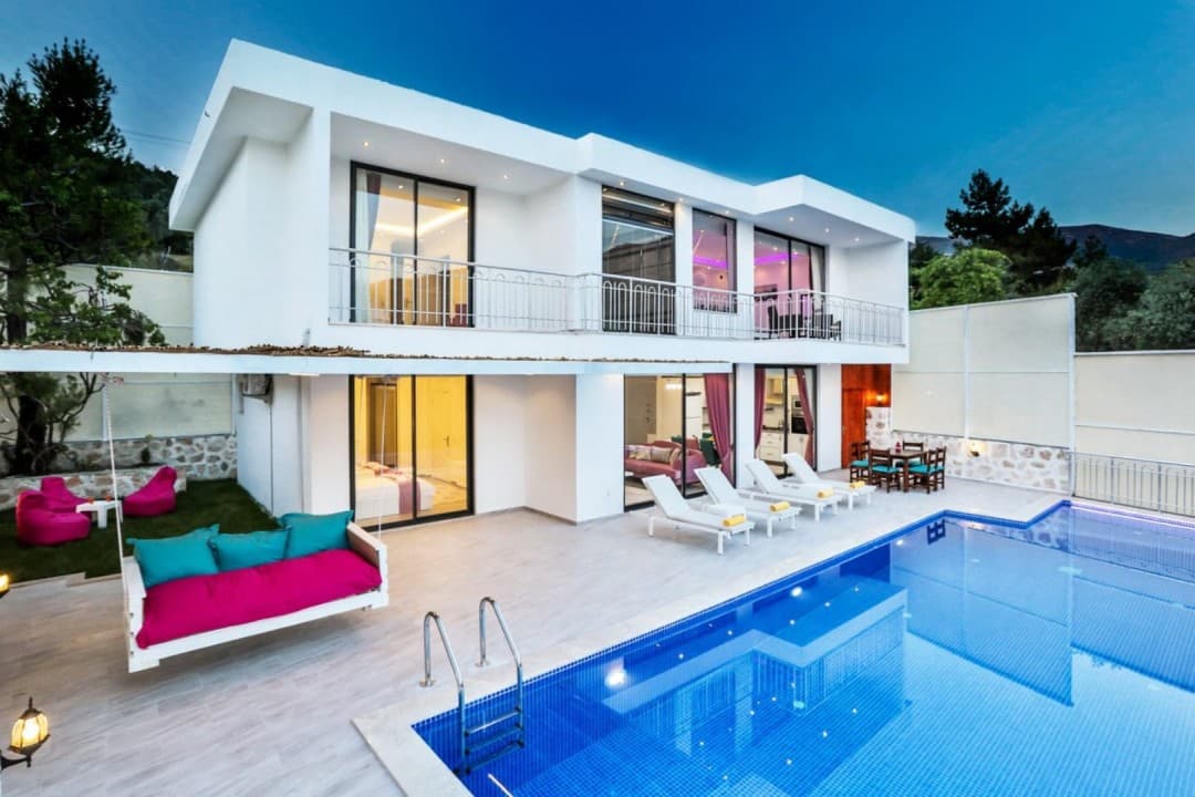 Akşam vakti aydınlatılmış, havuz başında şezlonglar, salıncak ve yemek masası bulunan iki katlı beyaz villa.
