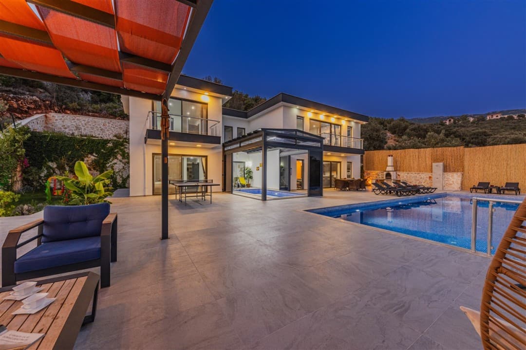 Akşam vaktinde aydınlatılmış, havuzlu, masa tenisi ve şezlong bulunan modern villanın dış görünüşü.