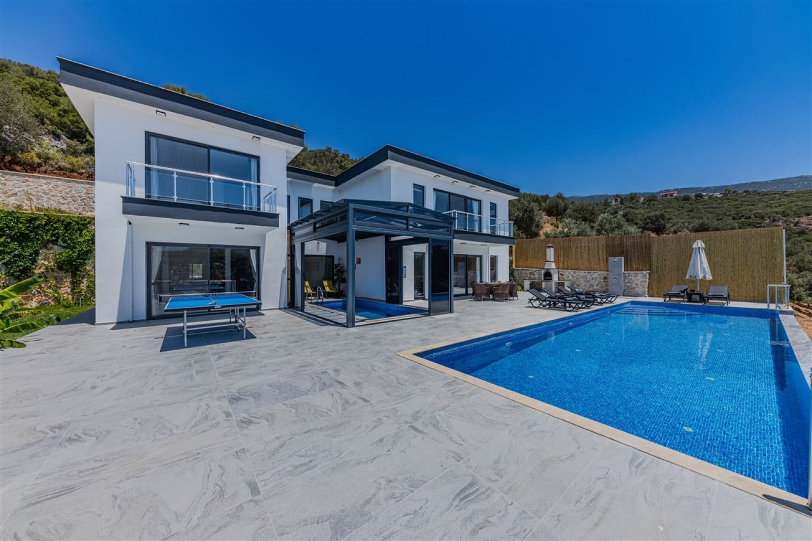 Geniş terasında yüzme havuzu, şezlonglar, masa tenisi ve kamelya bulunan beyaz villa dış cephesi.