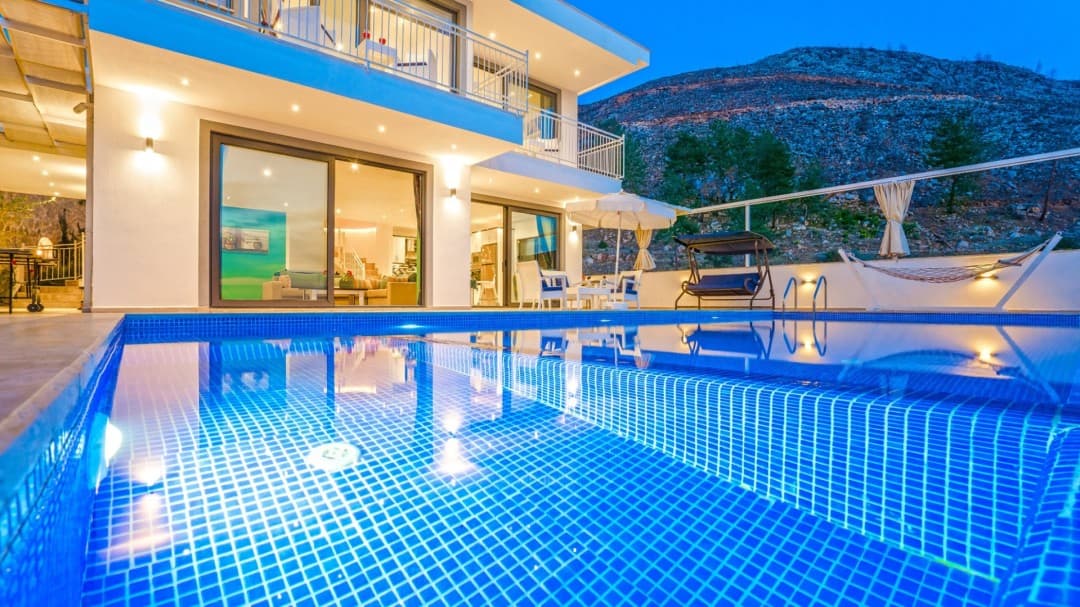 Akşam ışıklandırmalı havuz kenarında salıncak, şezlong ve şemsiye bulunan iki katlı modern villa.