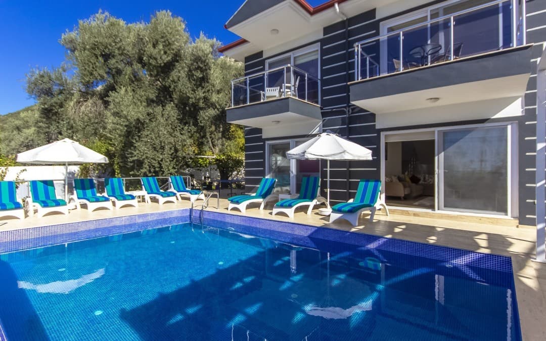 Mavi beyaz çizgili minderli şezlongların sıralandığı havuz ve iki katlı villa.