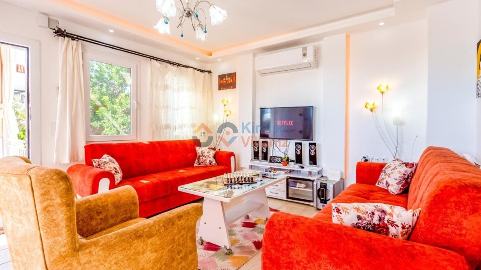 Kırmızı kanepeler, sarı berjer, sehpa ve televizyonun bulunduğu aydınlık iç mekan oturma odası.