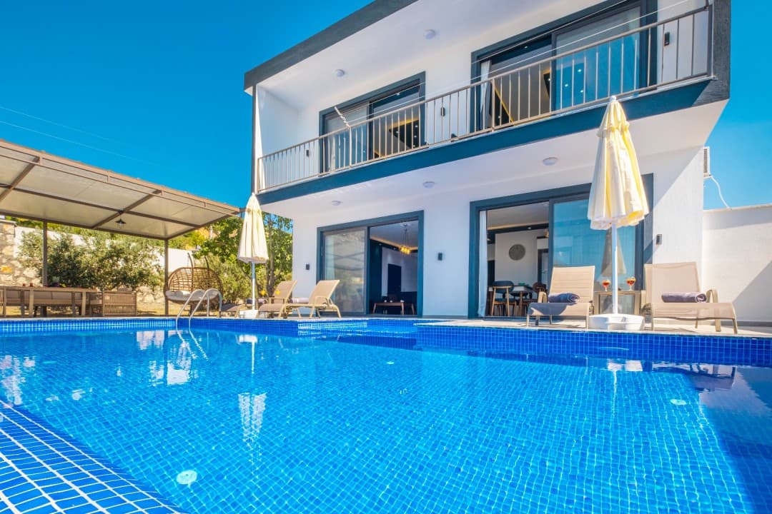 Mavi karolarla kaplı havuz kenarında şezlonglar ve iki katlı villa.