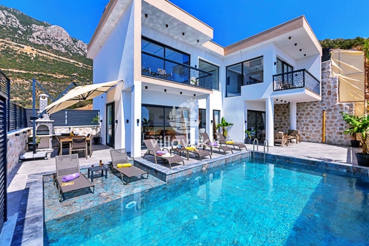 Dağ manzaralı havuz başında şezlonglar ve iki katlı beyaz villa.