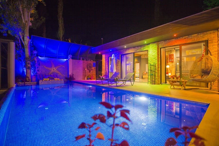 Gece aydınlatmalı havuz kenarında şezlonglar ve salıncak bulunan taş villa dış cephesi.