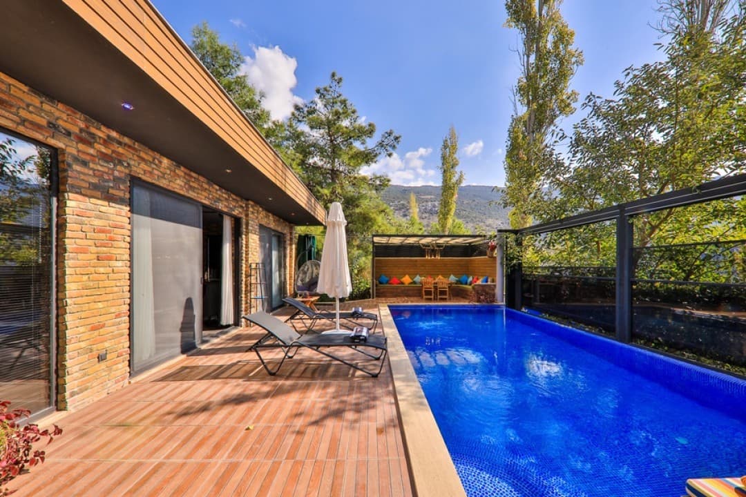 Tuğla duvarlı villa terasında şezlonglar, beyaz şemsiye ve yanında ağaçlarla çevrili özel yüzme havuzu.