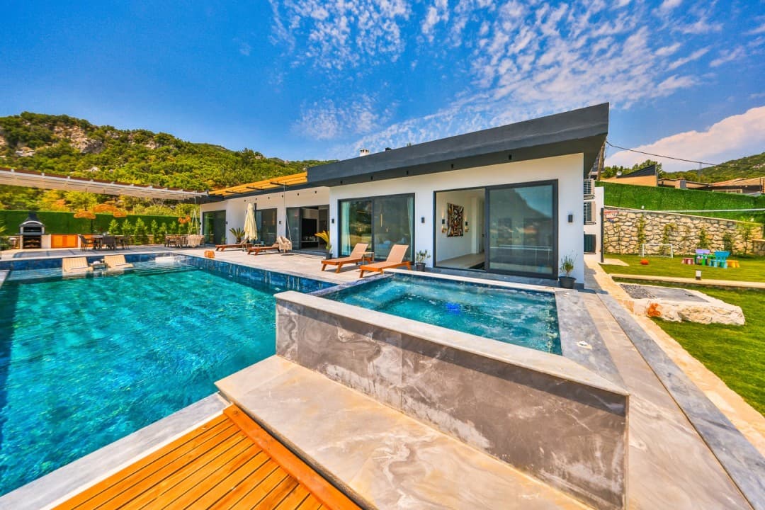 Ön planda havuz ve jakuzi, arkada tek katlı beyaz villa ve dağ manzarası.