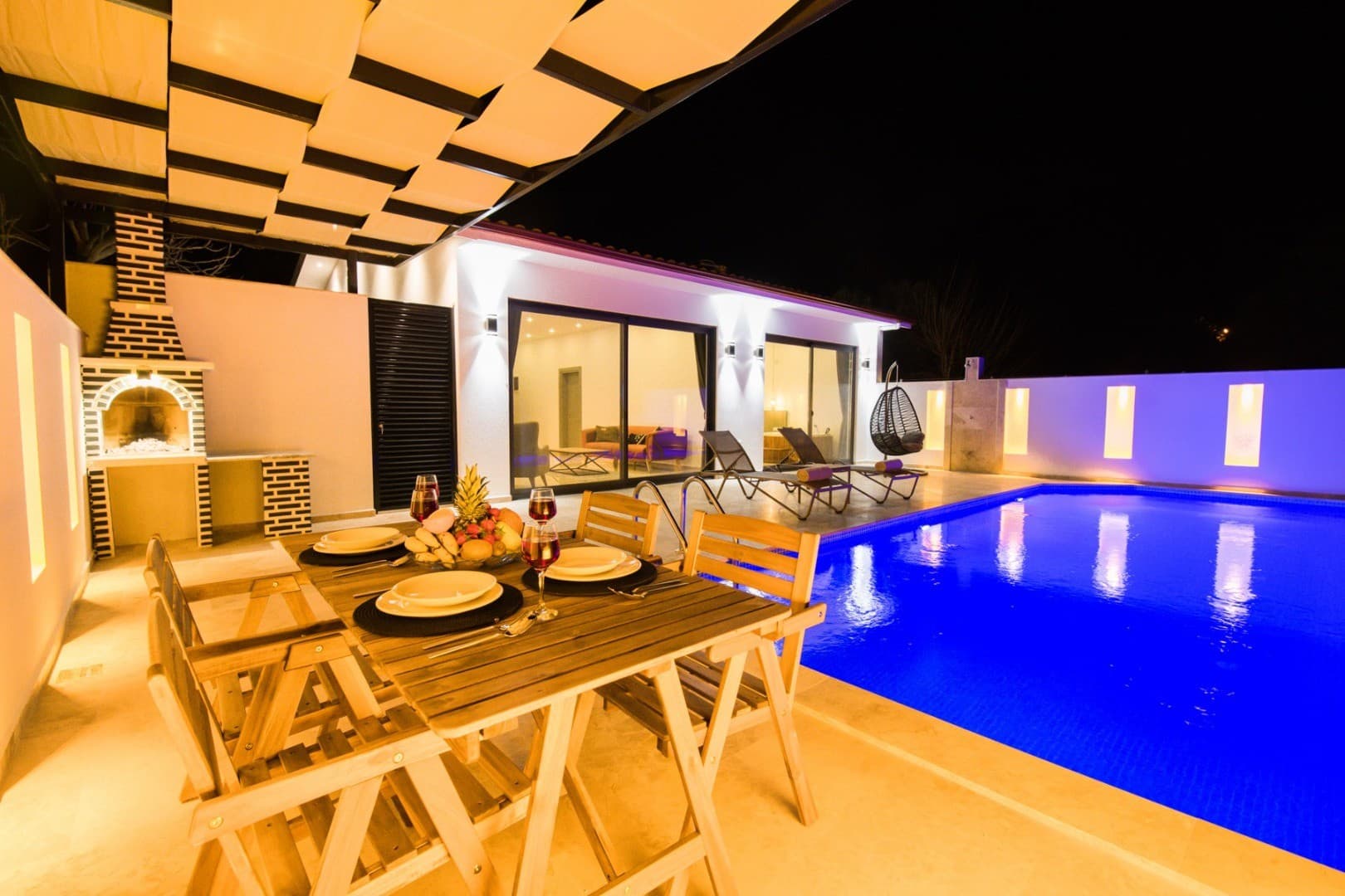Gece aydınlatmalı havuz kenarında ahşap yemek masası, barbekü alanı ve şezlongların bulunduğu villa terası.