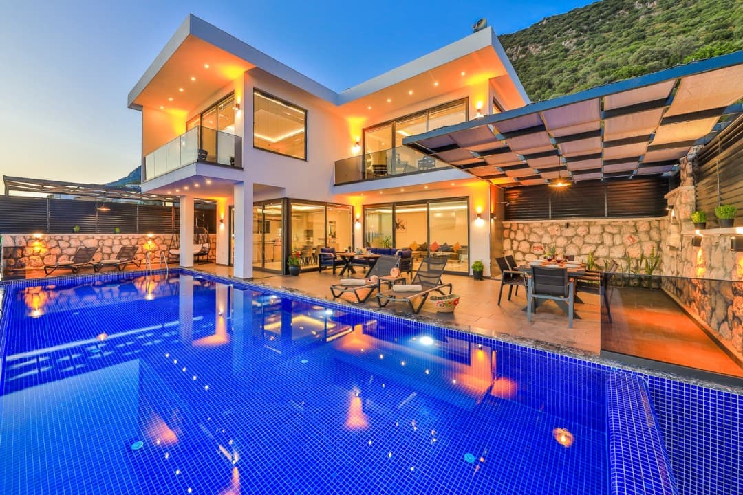 Akşam saatlerinde aydınlatılmış iki katlı modern villanın önünde havuz ve şezlonglar var.