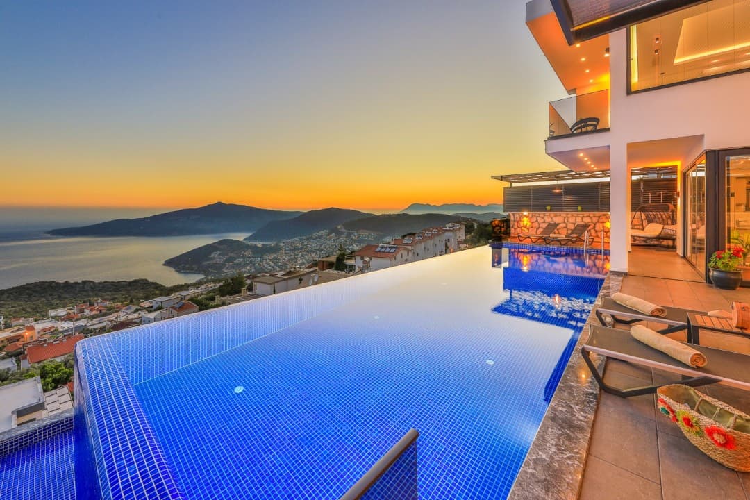 Gün batımında deniz ve dağ manzaralı sonsuzluk havuzu, havuz kenarında şezlonglar ve modern bir villa.