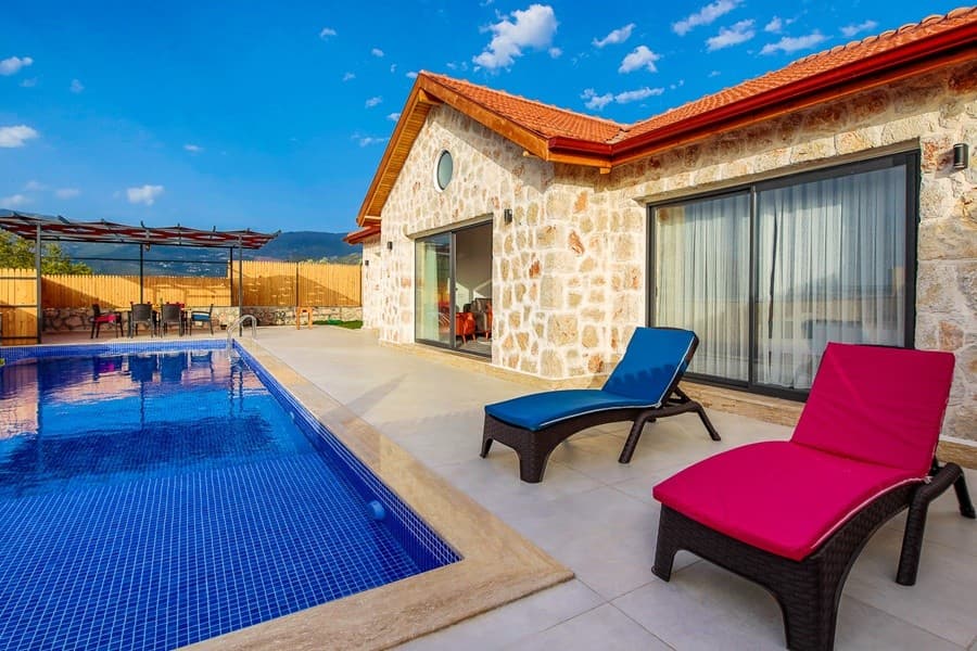 Mavi havuz kenarında pembe ve mavi şezlonglar olan taş cepheli villa.