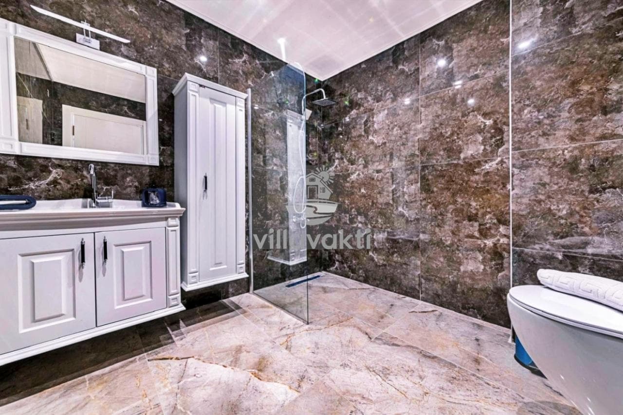 Kahverengi mermer kaplı banyoda duşakabin, beyaz lavabo ve klozet yer alıyor.