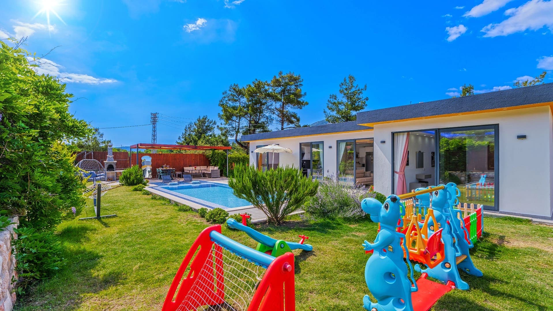 Bahçede yüzme havuzu, çocuk oyun alanı ve şezlonglar bulunan tek katlı beyaz villa.