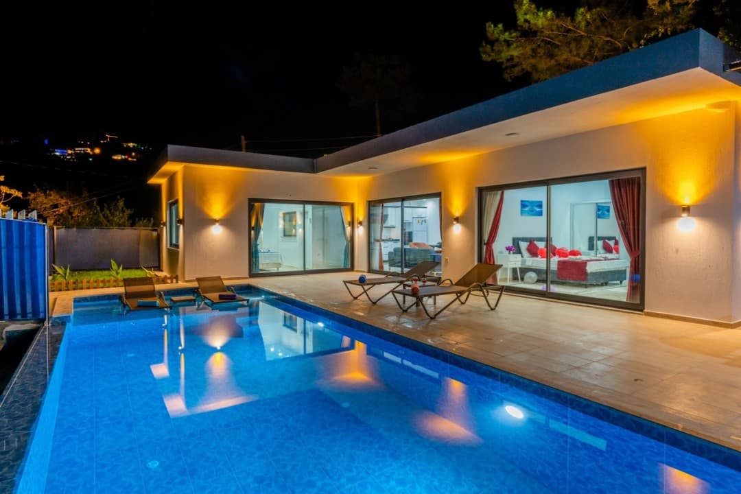 Gece aydınlatmalı havuz kenarında şezlonglar bulunan tek katlı villa.