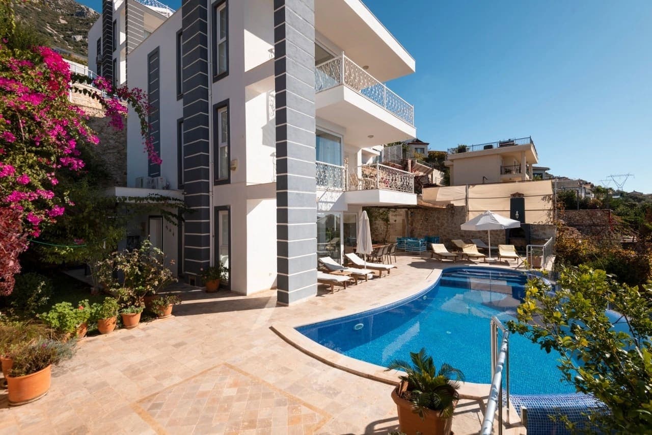 Önünde havuz, şezlonglar ve pembe çiçekli sarmaşık bulunan beyaz villa dış cephesi.