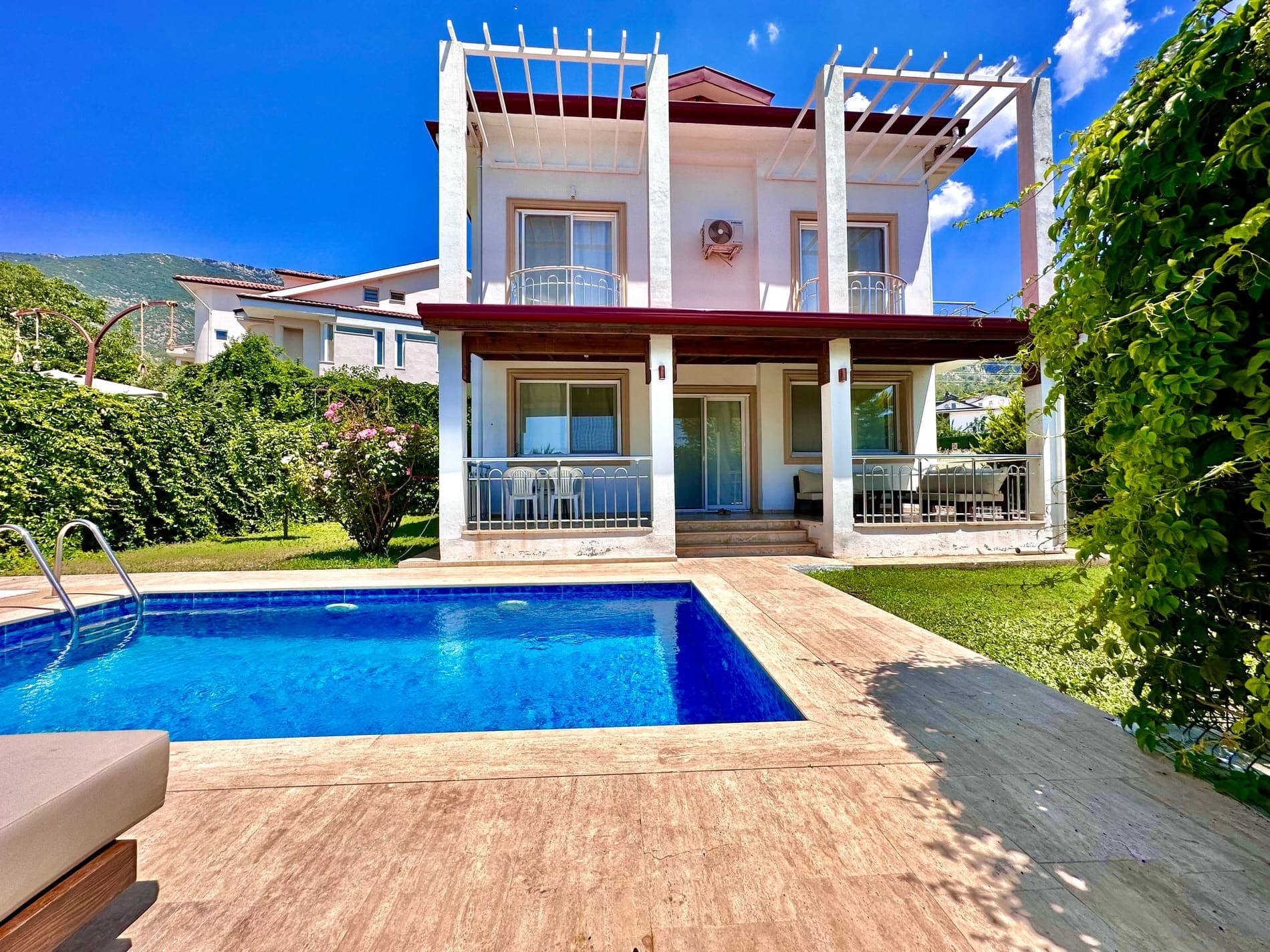 Mavi yüzme havuzu ve çim alan önünde iki katlı beyaz dış cepheli villa.