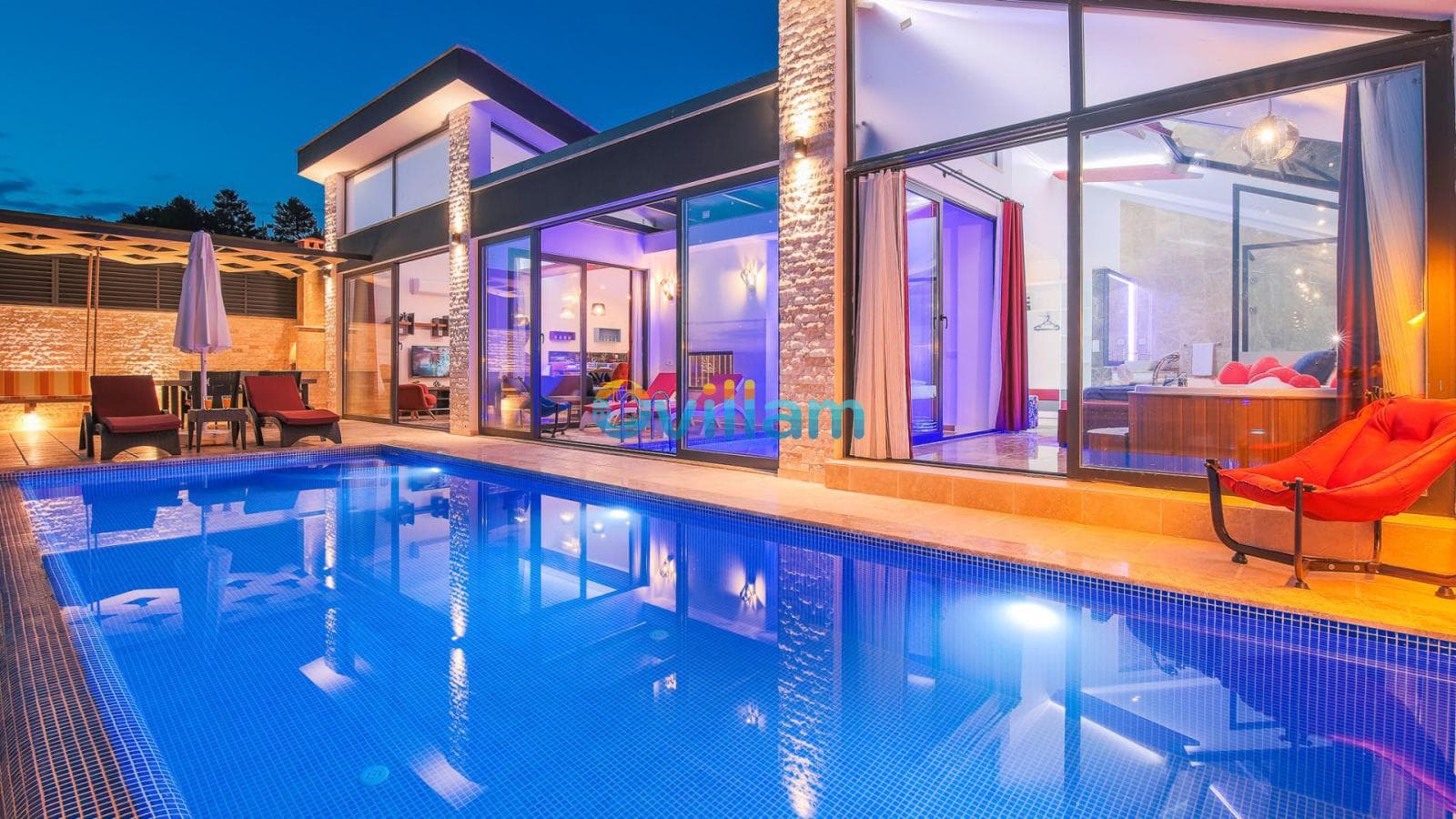 Akşamüstü mavi aydınlatmalı yüzme havuzu ve geniş camlı villa dış cephesi.
