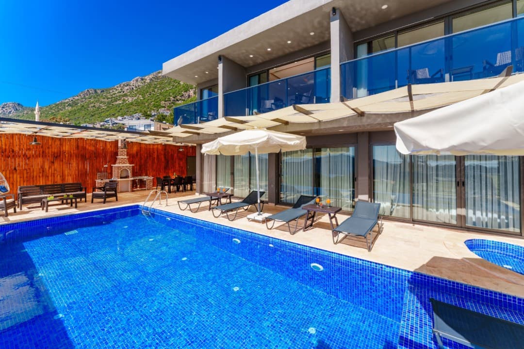 Mavi havuzlu, şezlonglu, güneş şemsiyeli ve taş barbekülü modern villanın dış görünümü.