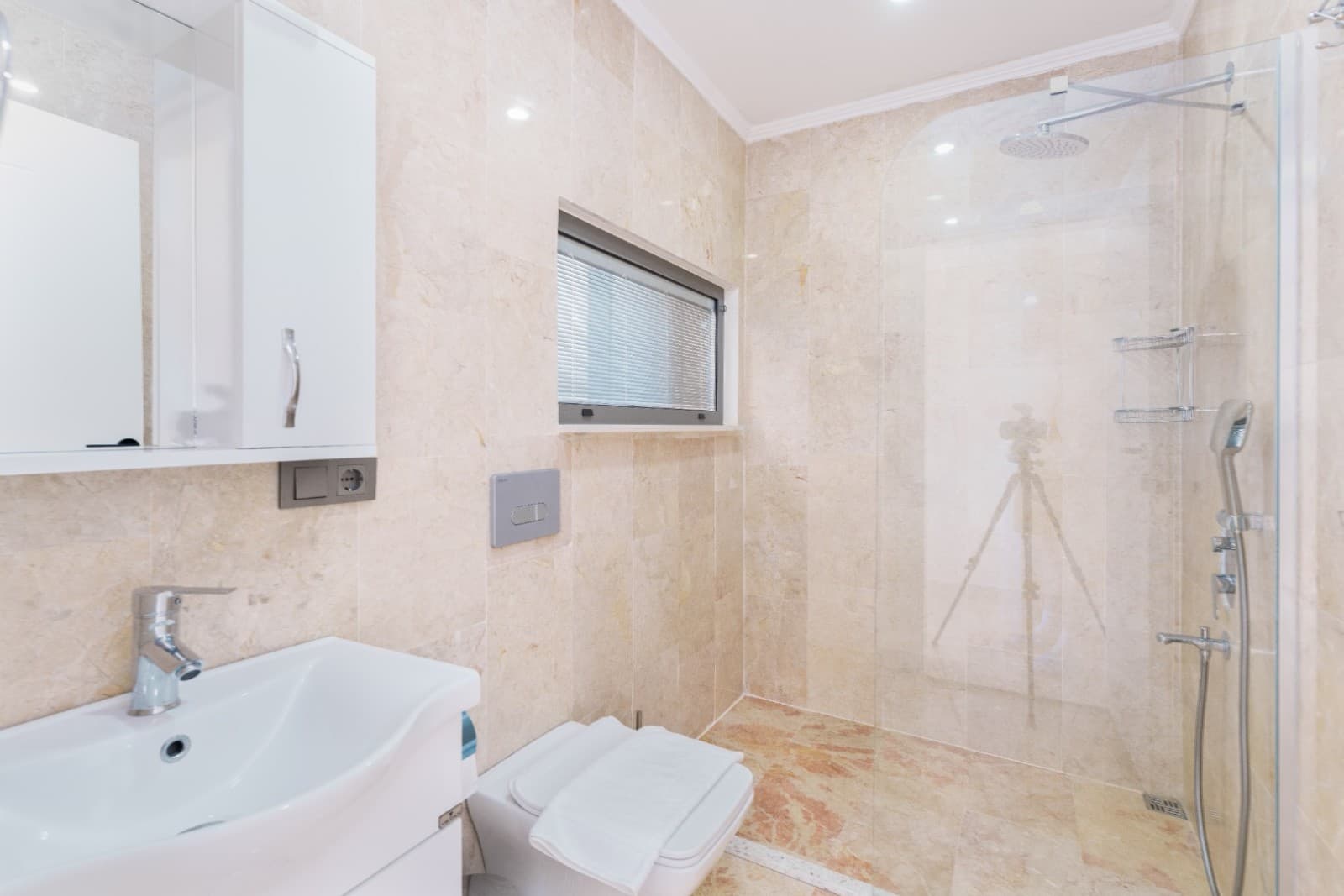 Bej mermer kaplı duşakabinli banyo, beyaz lavabo, asma klozet ve duş seti.