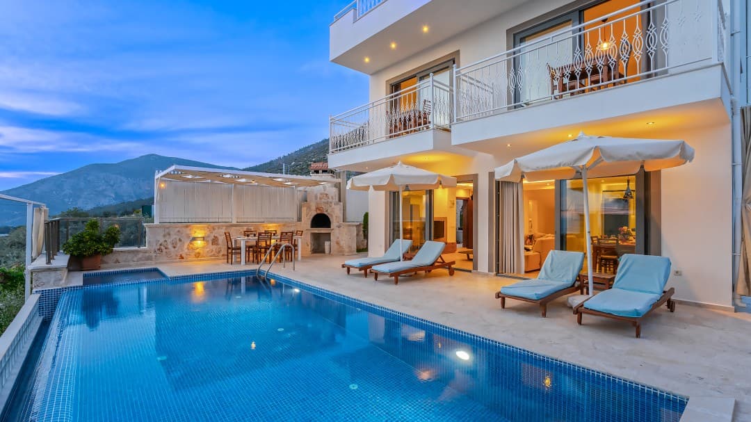 Akşam vakti havuz kenarında şezlonglar, şemsiyeler ve masalar bulunan iki katlı beyaz villa.