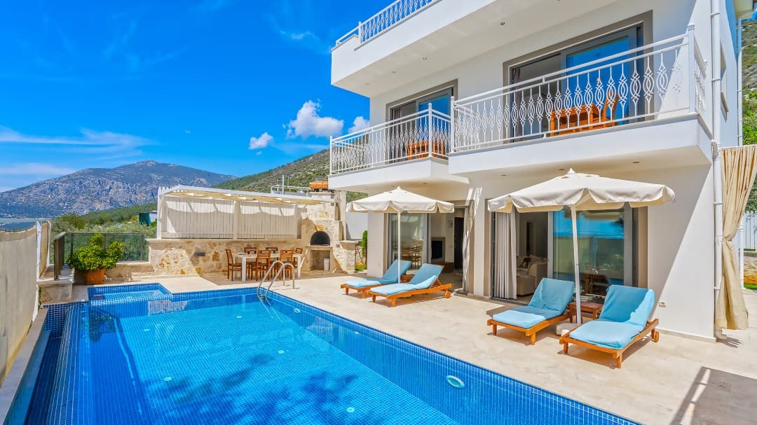 Mavi havuzlu, şezlonglu, taş barbekülü ve iki katlı beyaz dış cepheli villa.