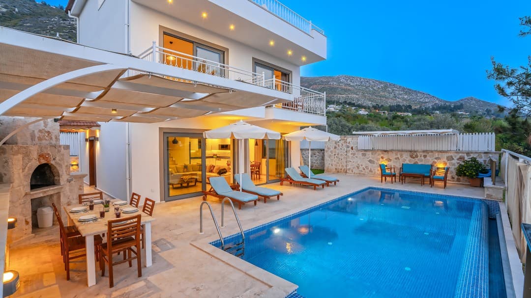 Havuz kenarında şezlonglar, yemek masası ve arkada dağ manzaralı iki katlı beyaz villa.