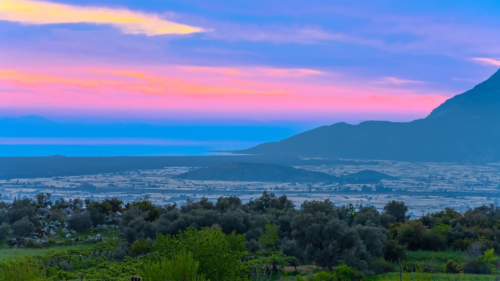 Pembe ve mavi tonlarında gün batımı altında ovalık arazi ve dağ manzarası.