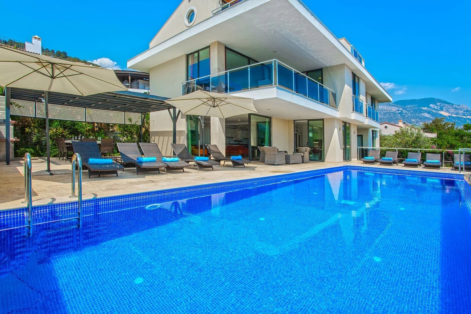 Geniş mavi yüzme havuzu ve önünde şezlonglar bulunan modern villanın dış cephesi.