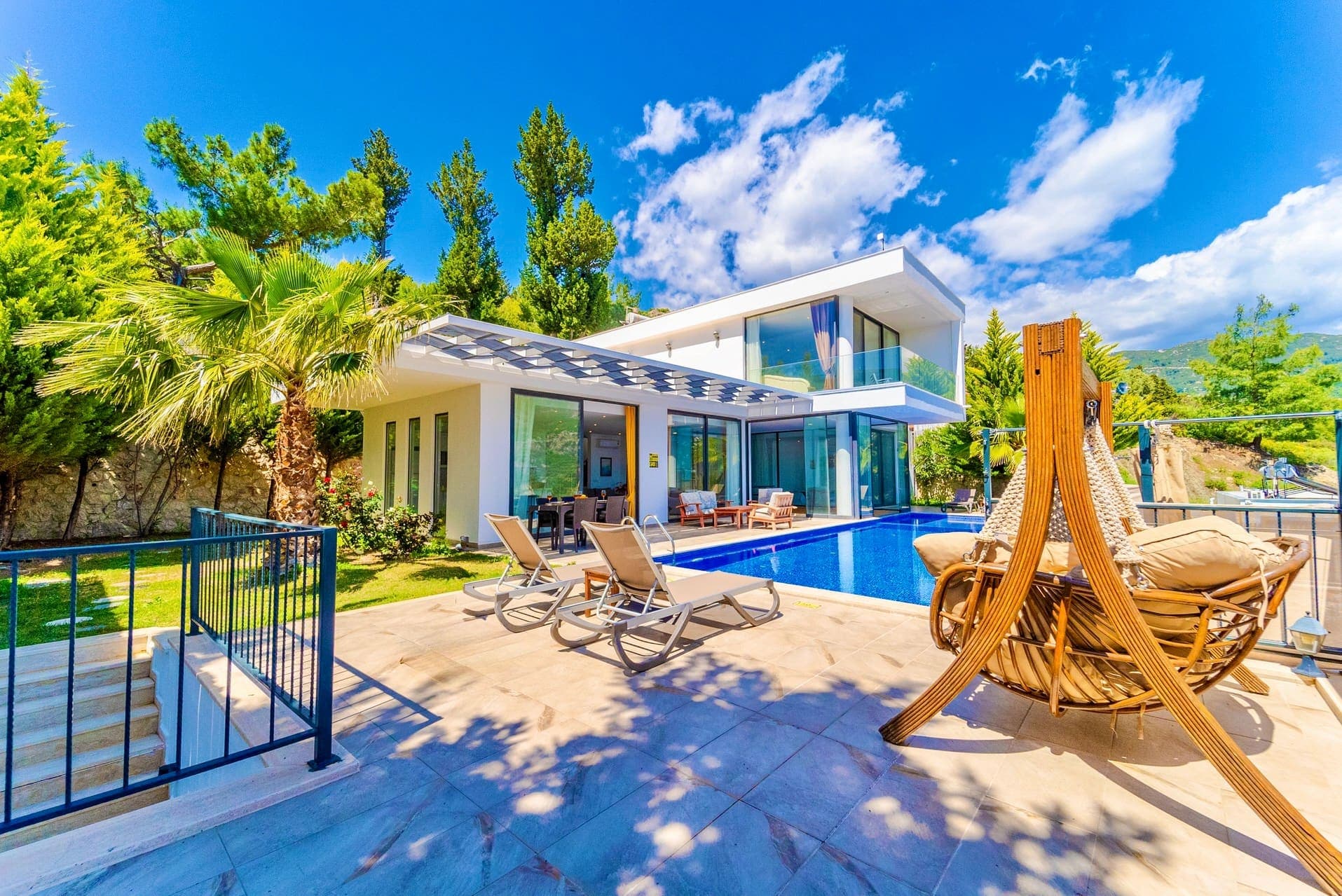 Güneşli havuz terasında şezlonglar, salıncak ve arkada iki katlı modern villa.