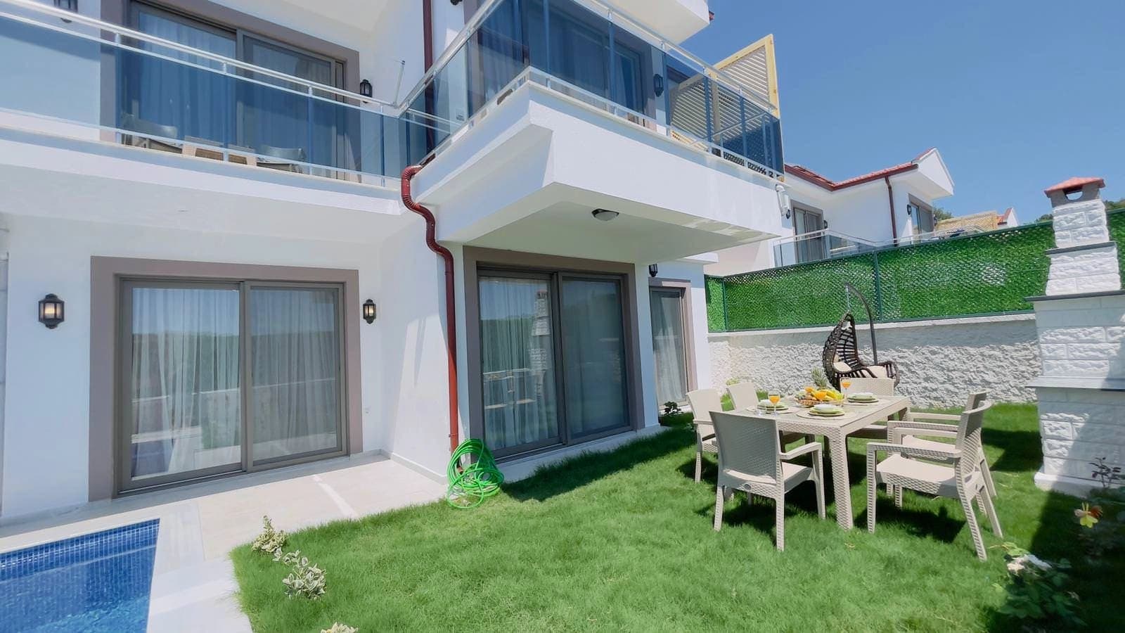 Beyaz dış cepheli villanın bahçesinde yemek masası ve şömine yer alıyor.