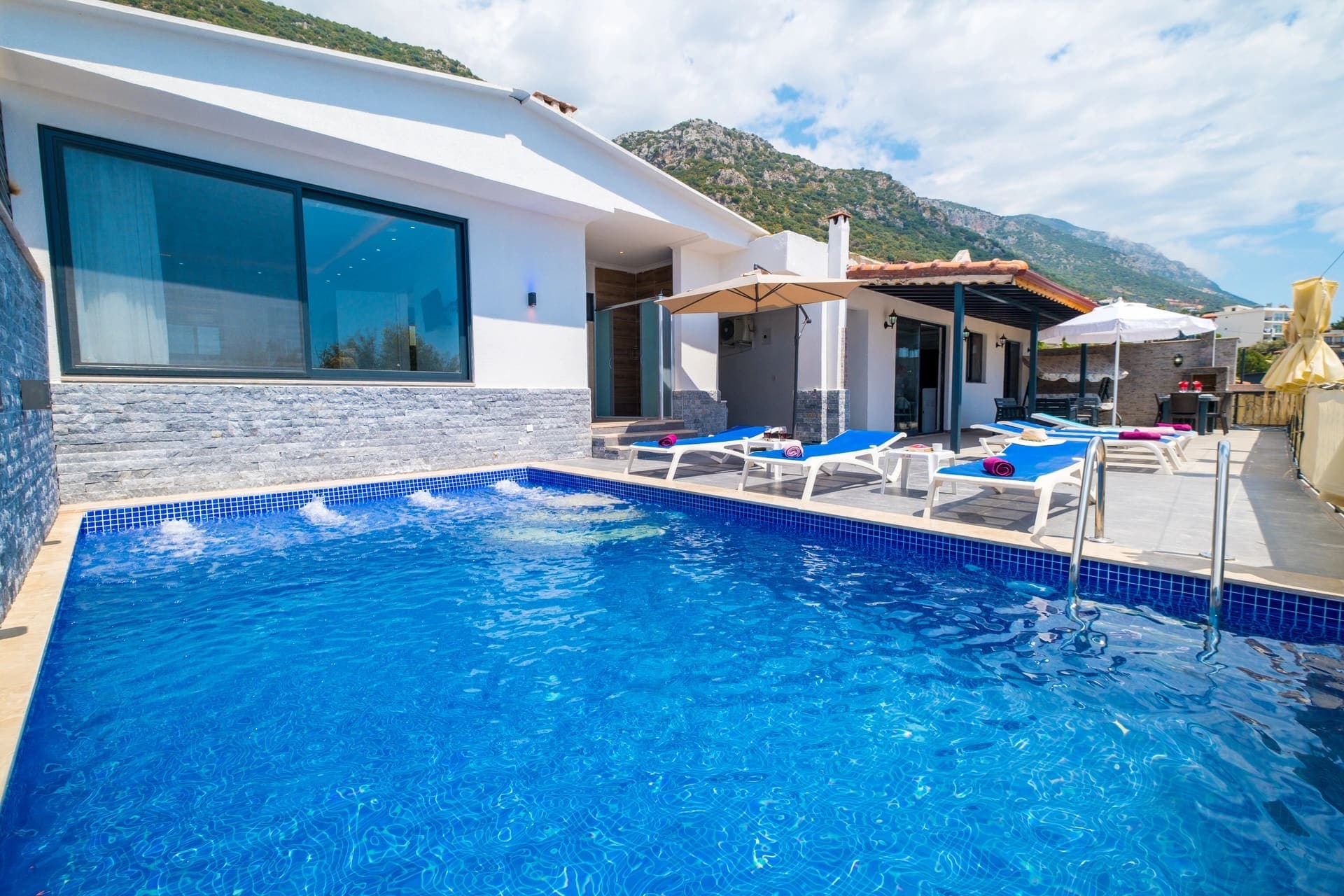 Dağ manzaralı havuz kenarında şezlonglar ve şemsiyeler bulunan beyaz villa dış cephesi.