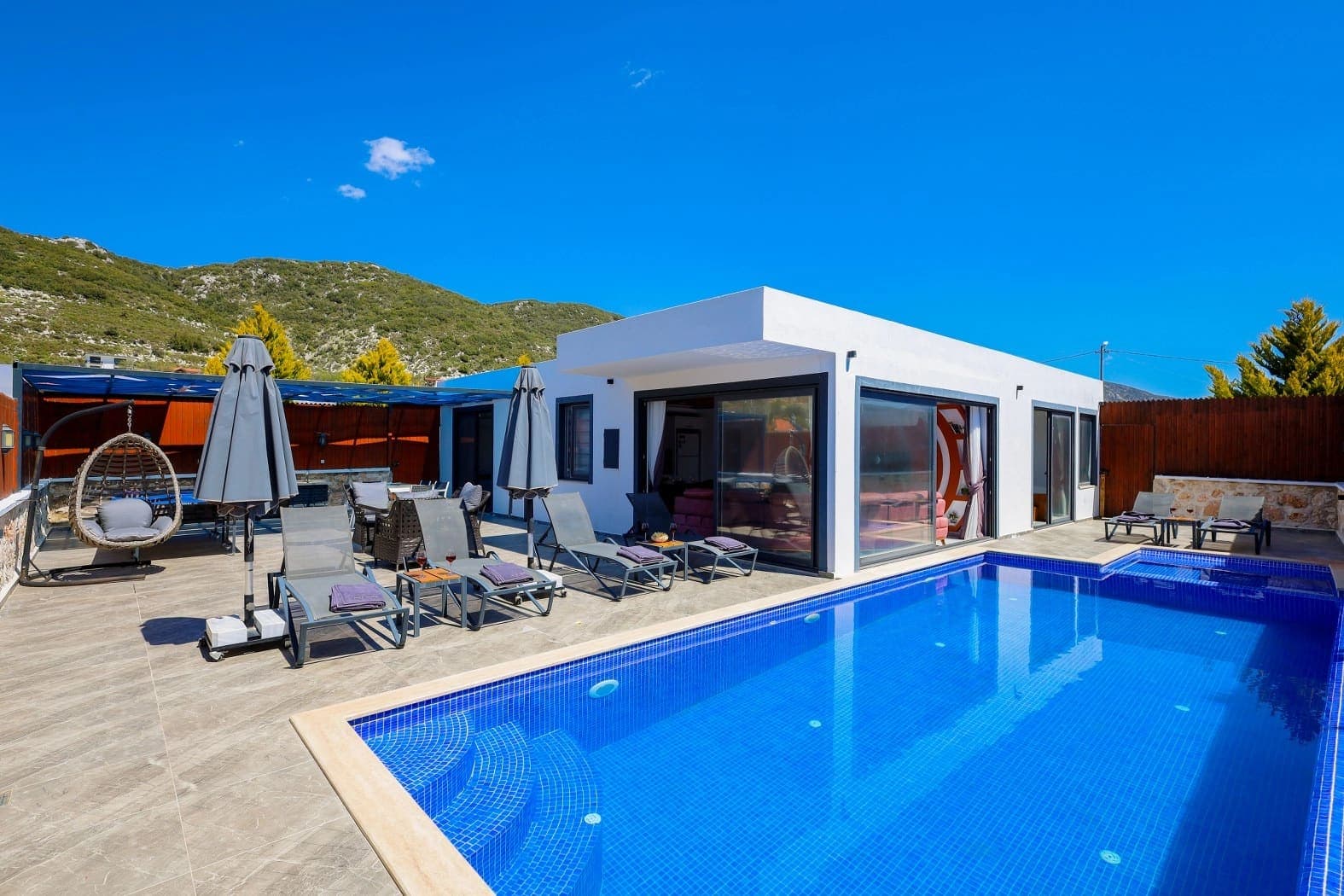 Mavi havuzun kenarında şezlonglar, şemsiyeler ve arkada dağ manzarası olan villa dış cephesi.