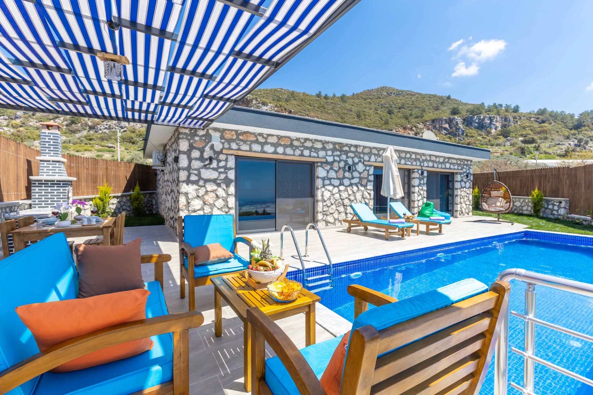Taş duvarlı villa önünde mavi havuz, şezlonglar ve oturma grubu.