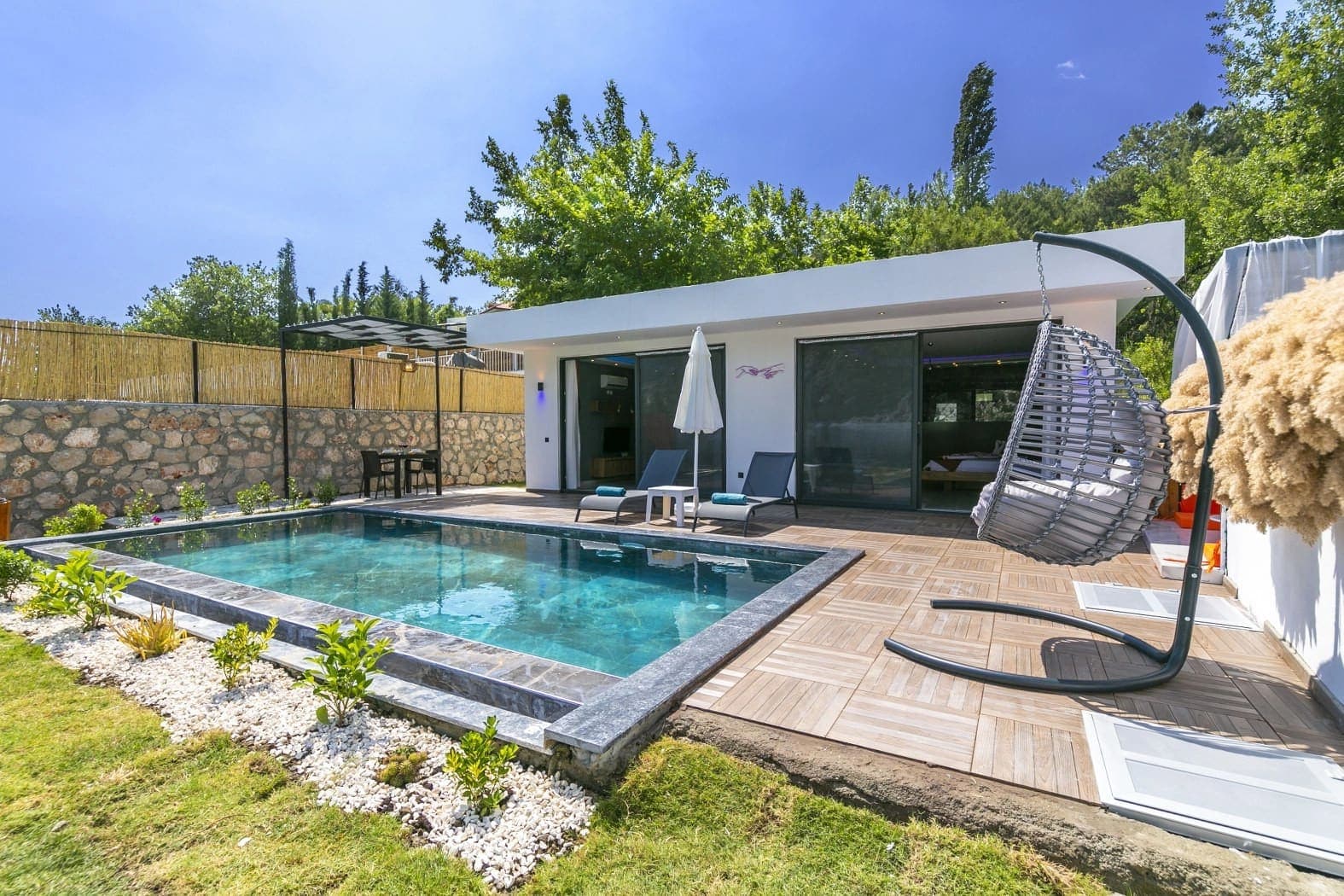 Orman manzaralı havuz kenarında şezlonglar, salıncak ve tek katlı villa.