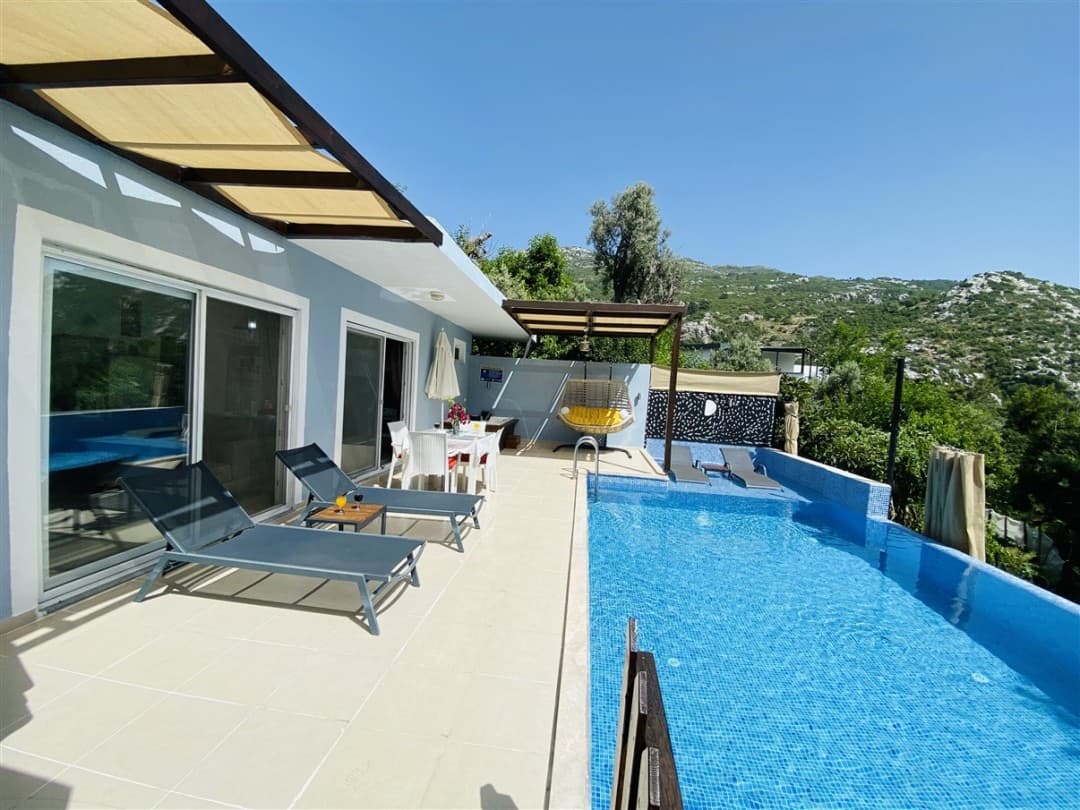 Mavi havuzlu, şezlonglu, yemek masalı ve asma salıncaklı villa terası ile dağ manzarası.