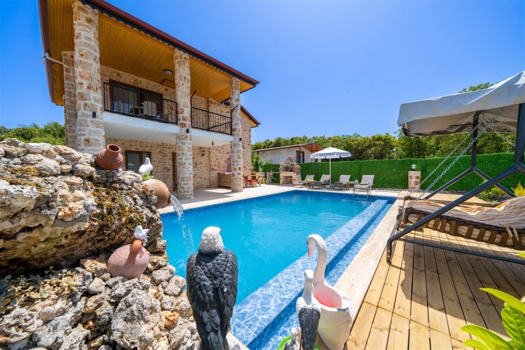 Taş kaplama iki katlı villa, önünde yüzme havuzu, havuz kenarında heykeller ve ahşap zeminli bir salıncak.