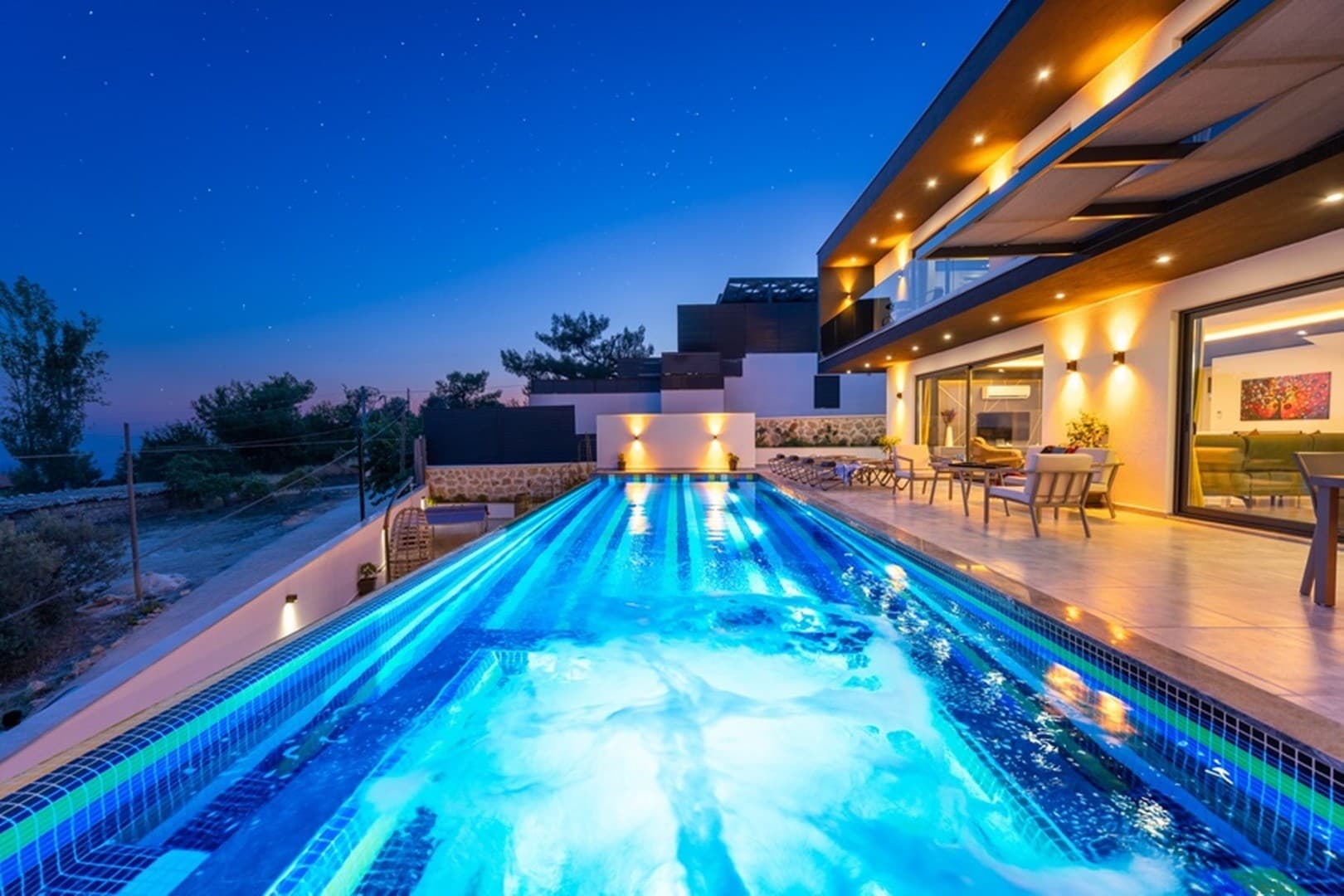 Akşam vakti aydınlatılmış uzun havuzu ve oturma grubu olan modern villanın dış görünümü.