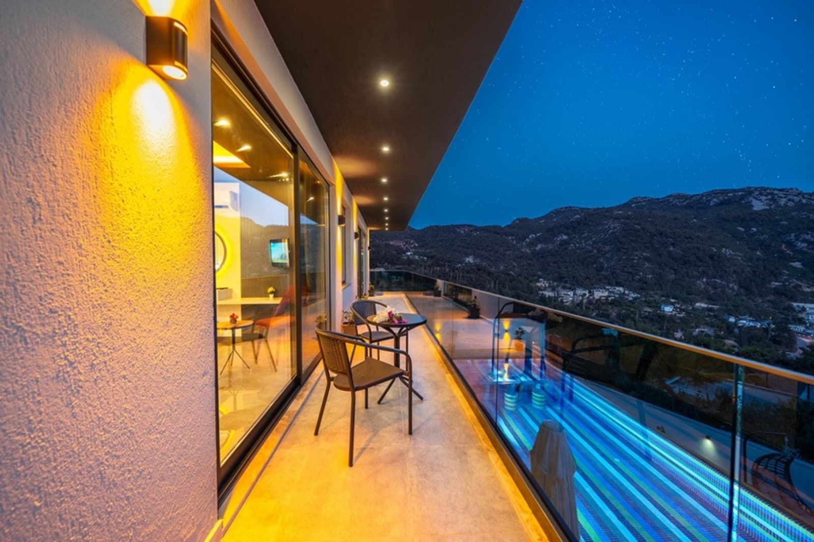 Gece vaktinde ışıklandırılmış, havuz ve dağ manzaralı geniş balkon.