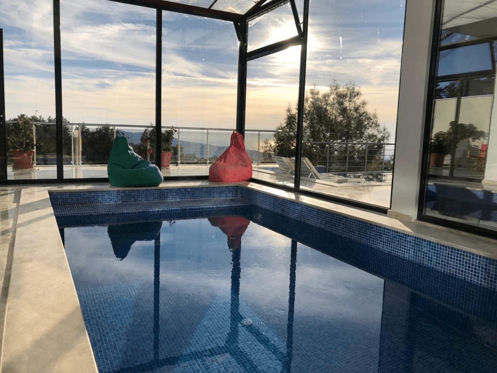 Camla kapalı havuz alanında renkli minderler ve gün batımı manzarası.